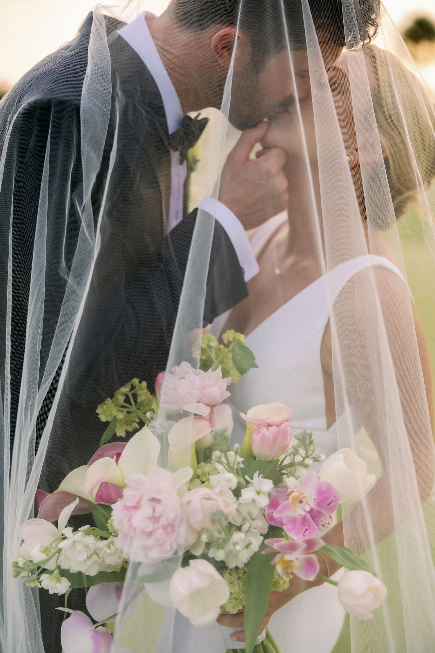 Bride and groom sharing a romantic moment under a flowing veil, with a lush pink and white floral bouquet.