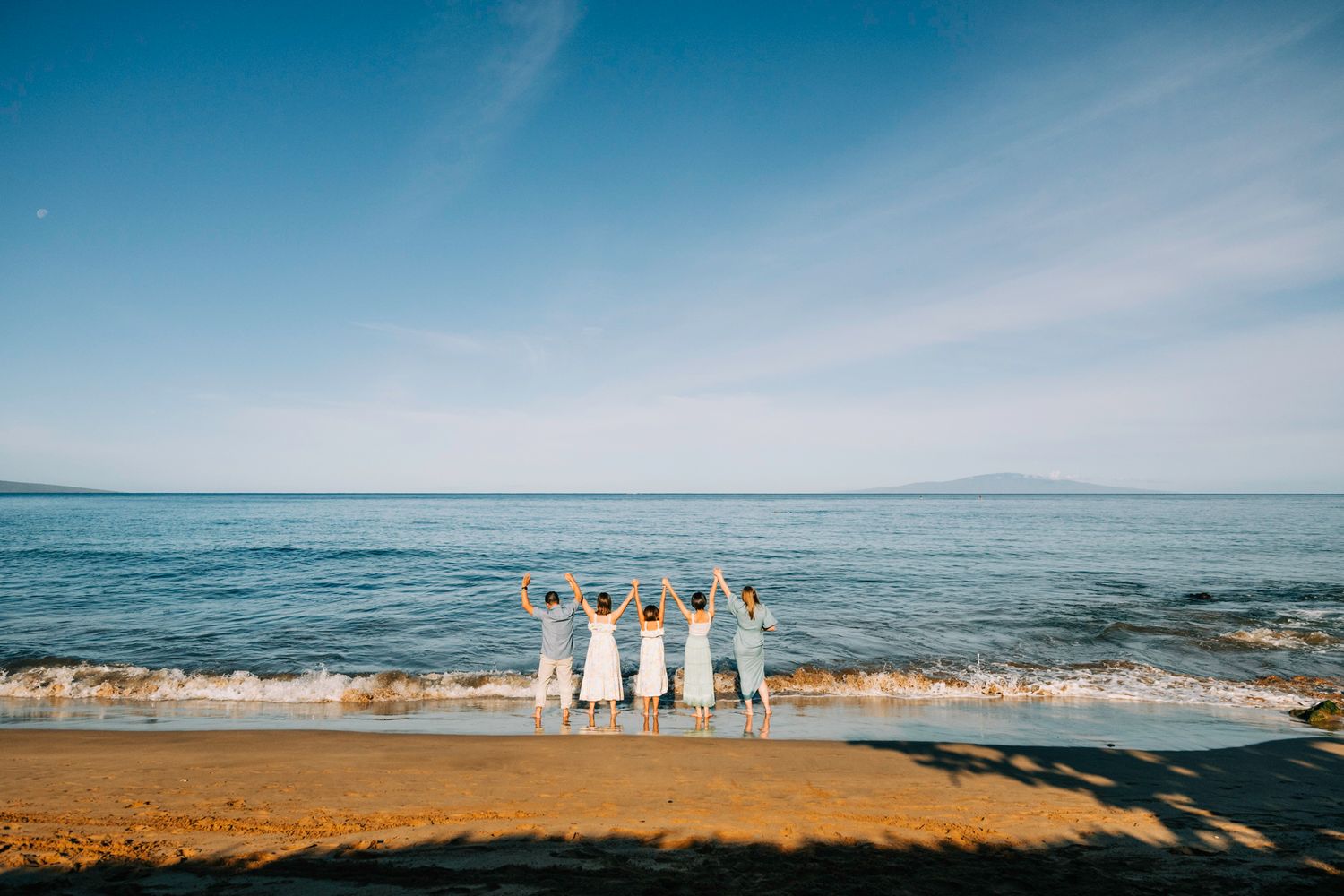 Four women in white dresses celebrating with raised arms at the edge of a sunny beach.