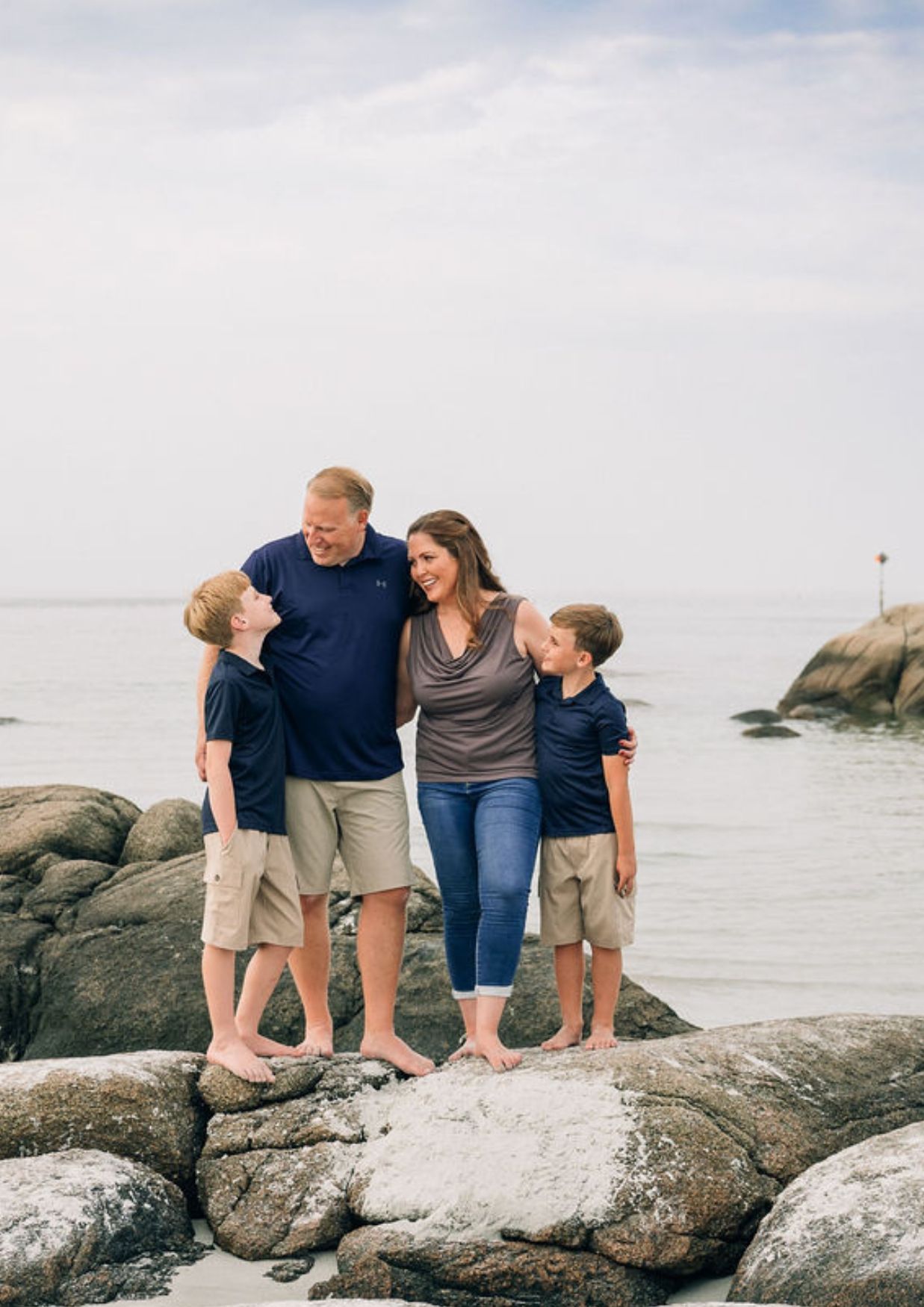 Family of four smiling together on rocky beach, two boys and parents in navy and khaki, ocean backdrop.