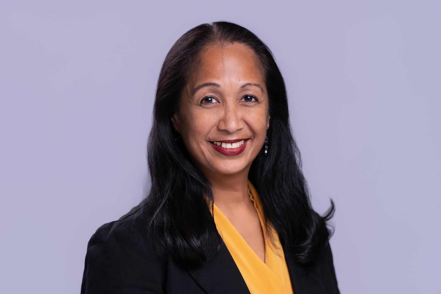 Professional headshot of a smiling woman in a black blazer and yellow top against a gray background.