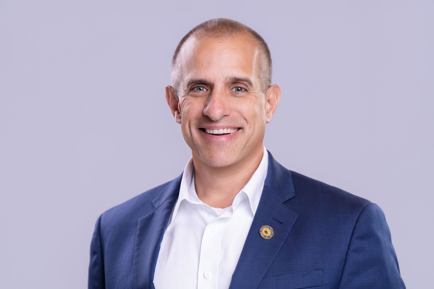 Professional headshot of a smiling middle-aged man in a navy blue blazer and white shirt against a gray background.