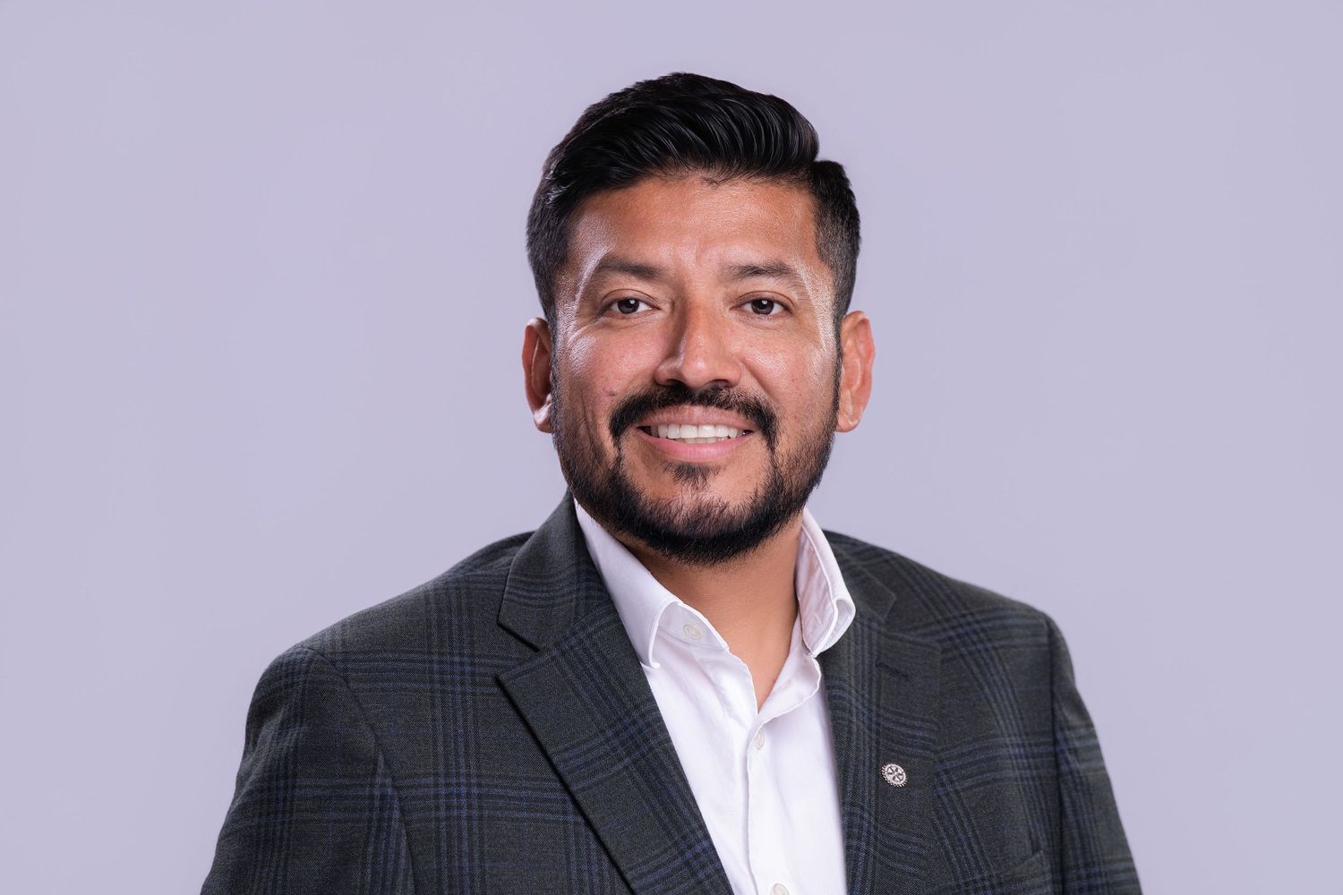 Professional headshot of a smiling Hispanic businessman in a gray blazer against a light gray background.