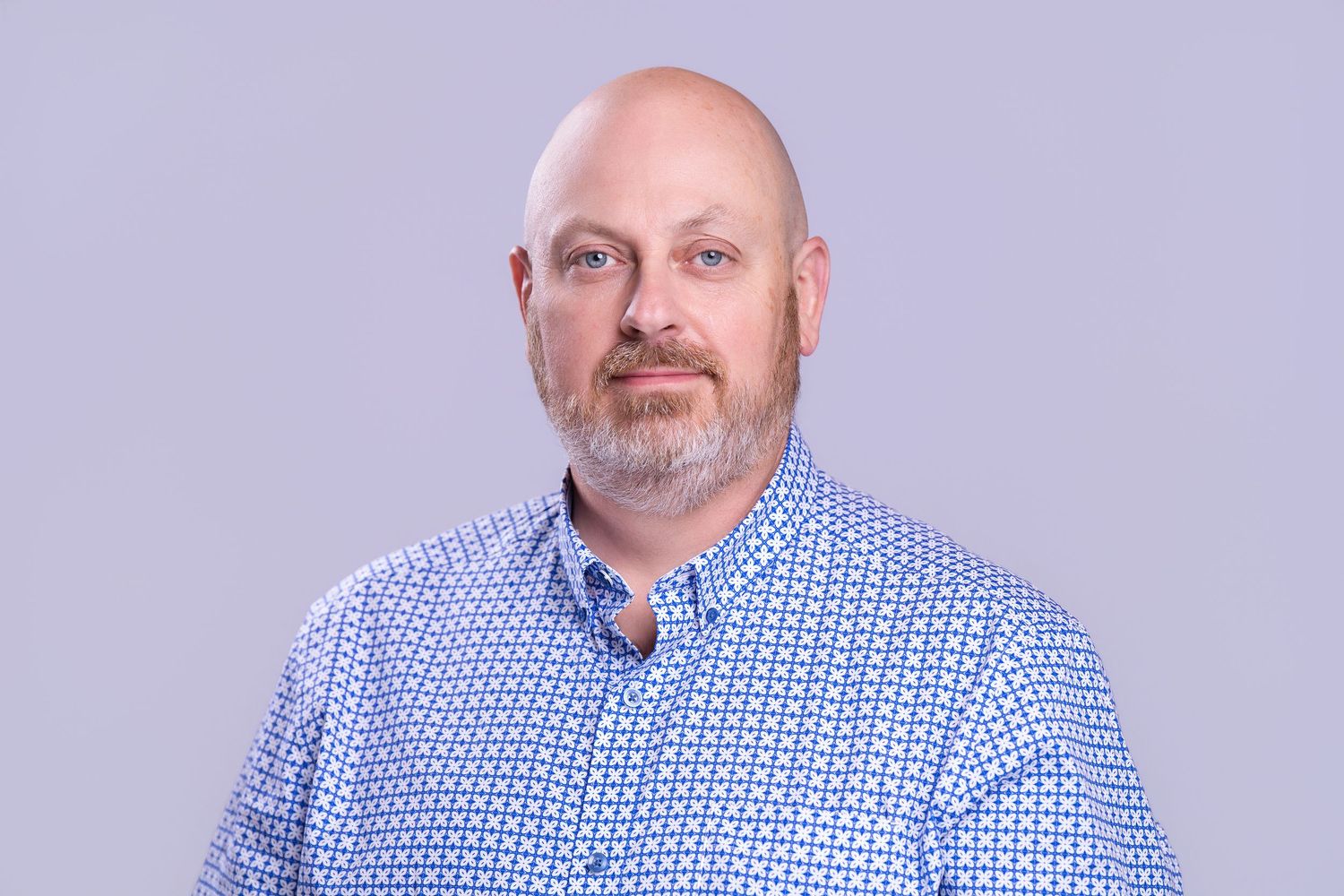 Professional headshot of a bald man with a gray beard wearing a blue patterned shirt against a gray background.