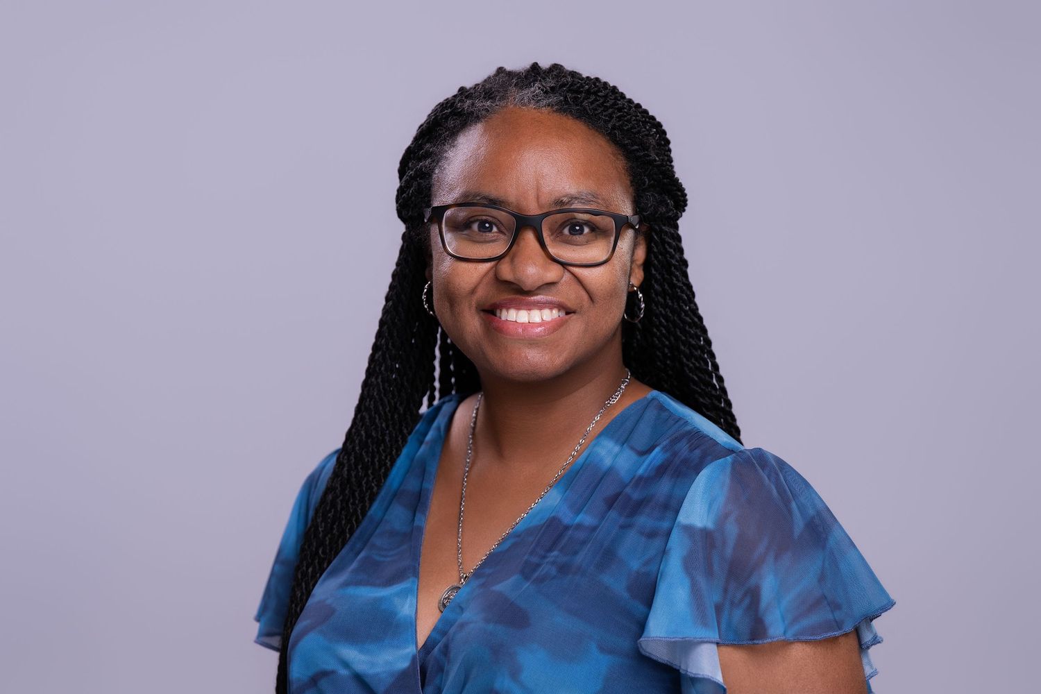 Smiling young woman with braids and glasses wearing a blue floral top, professional headshot on gray background.