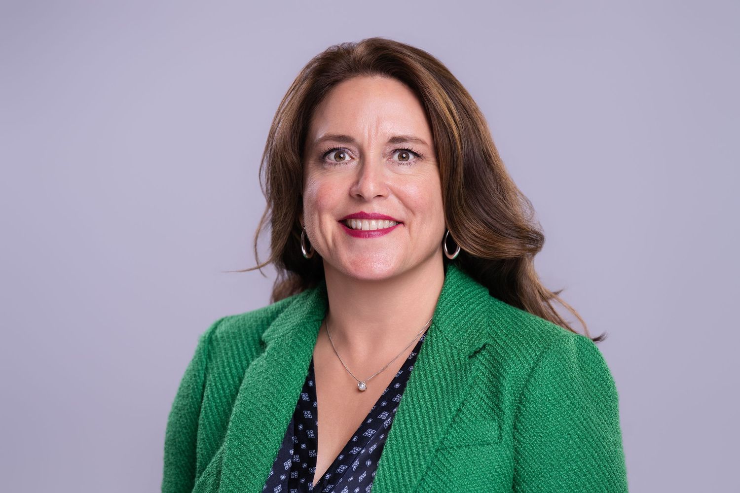 Professional woman in green blazer and floral blouse smiling confidently against gray background.