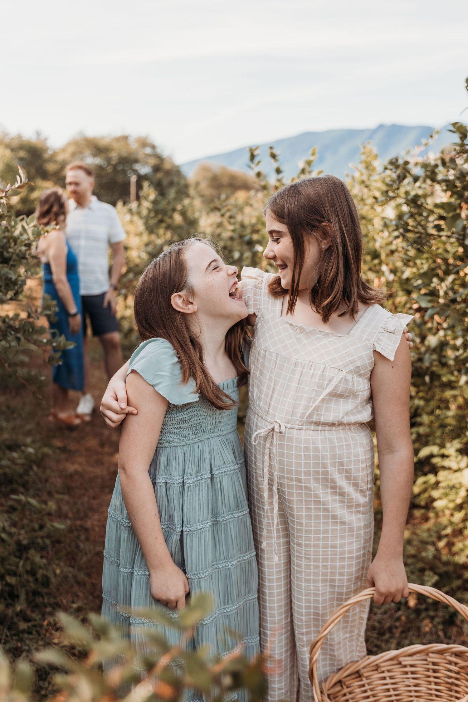 Two young girls laughing and hugging in an orchard, holding a basket, with family in the background.