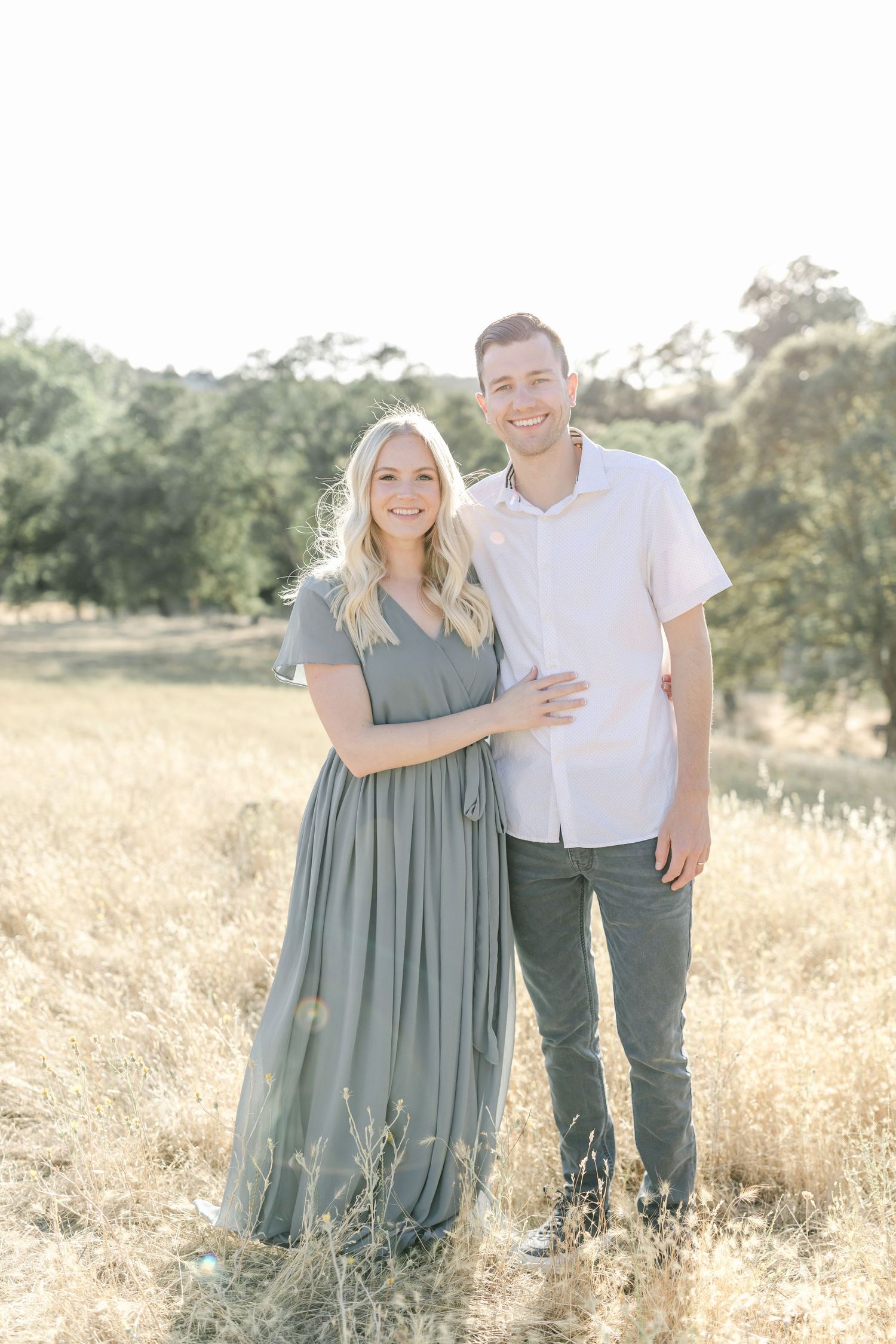 A smiling couple poses in a golden field, woman in sage maxi dress, man in white shirt and gray pants.