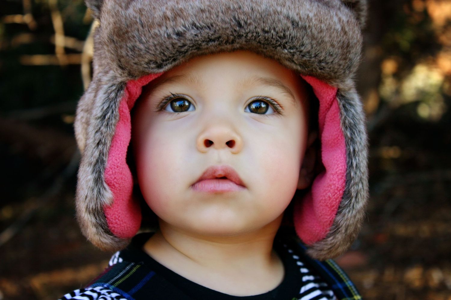 A toddler with wide eyes wears a fuzzy fur-trimmed winter hat with pink lining, standing outdoors against a blurred woodland background.