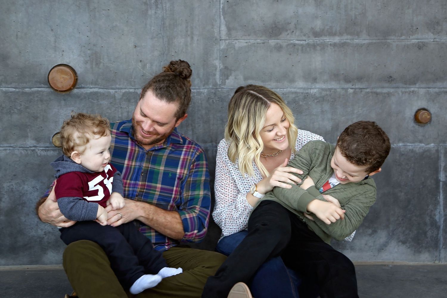 Smiling family of four sits against a concrete wall as a bearded dad and blonde mom hold their toddler and young son.