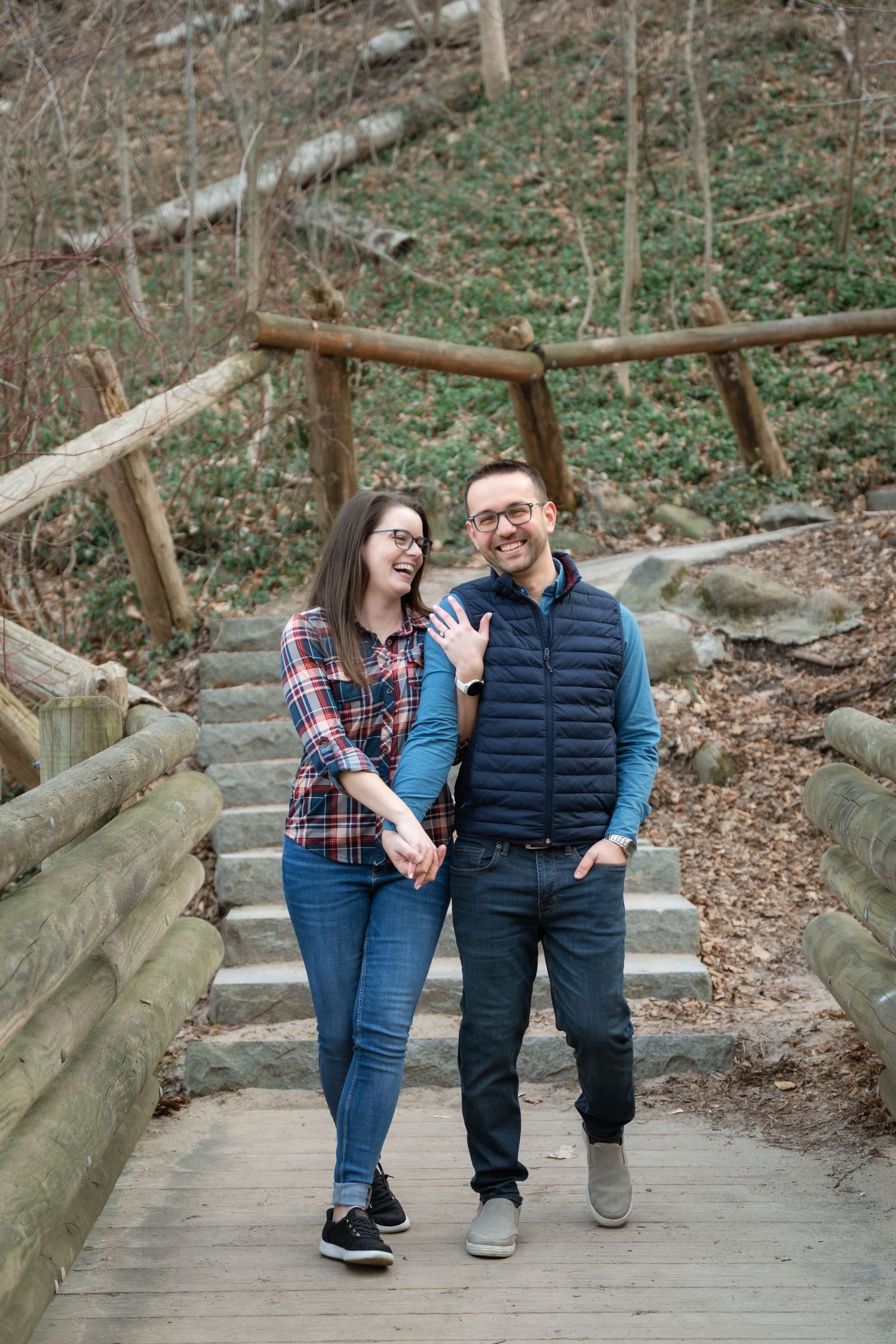 Couple embracing on a wooded trail in autumn, woman in plaid scarf and jeans, man in blue vest, sharing a tender moment.