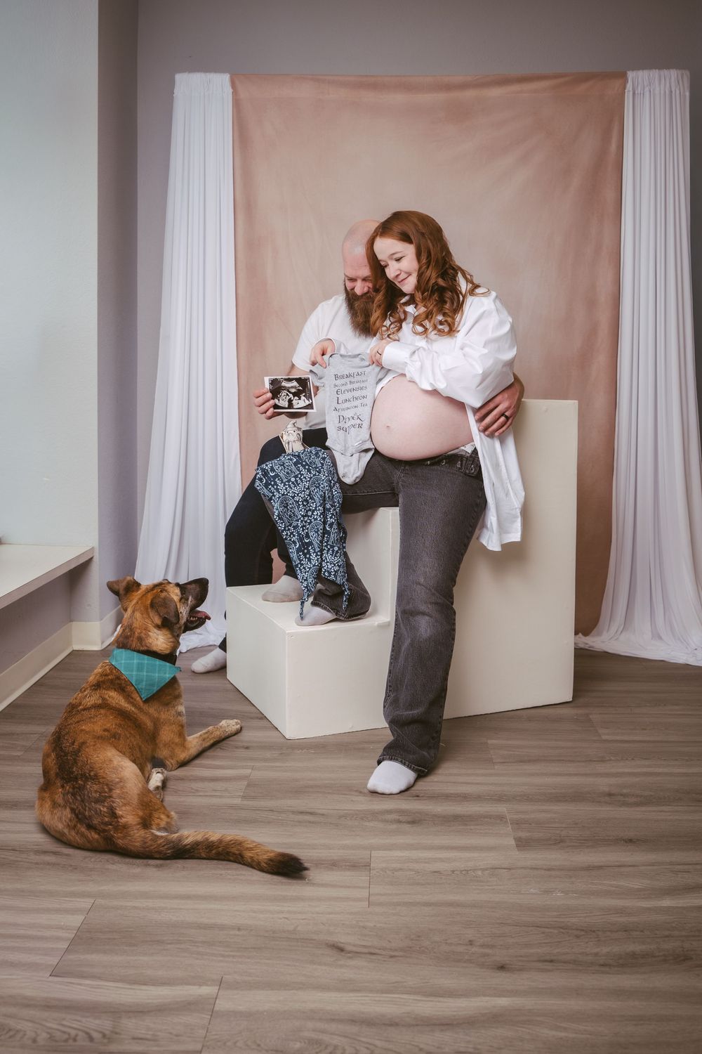 Pregnant couple smiling at ultrasound photos with their brown dog wearing a teal bandana in a studio.