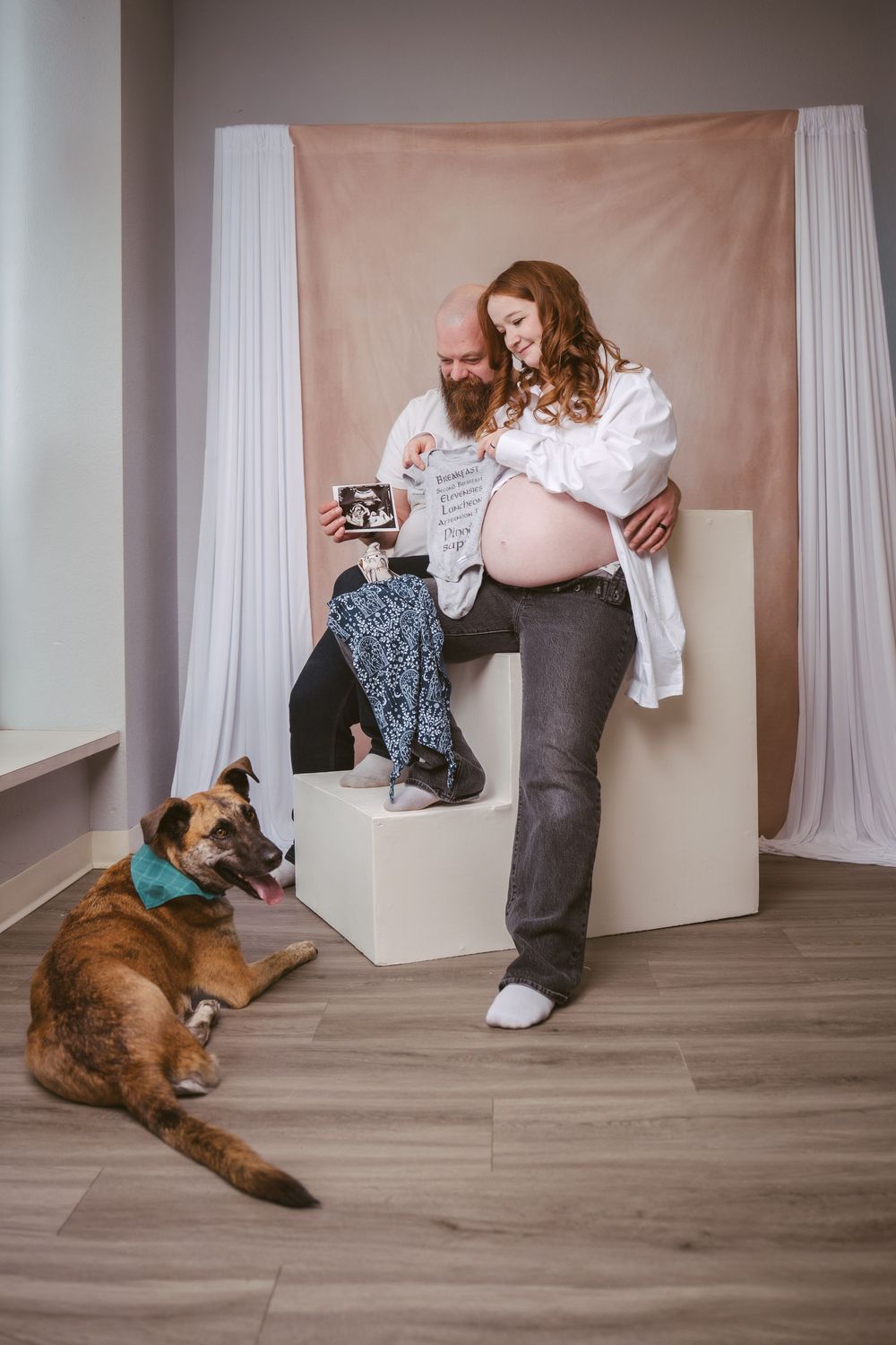 Pregnant couple with dog reviewing ultrasound photos and baby clothes during a maternity photo session.
