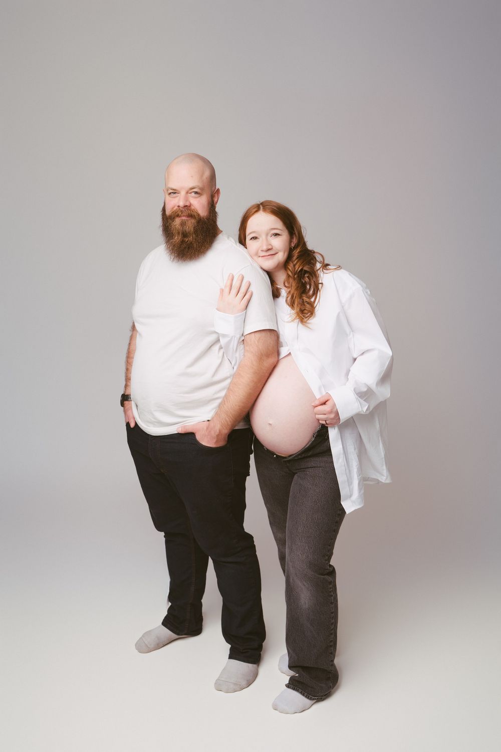 Pregnant woman with red hair poses with bearded man in white tee, both smiling in a studio setting.