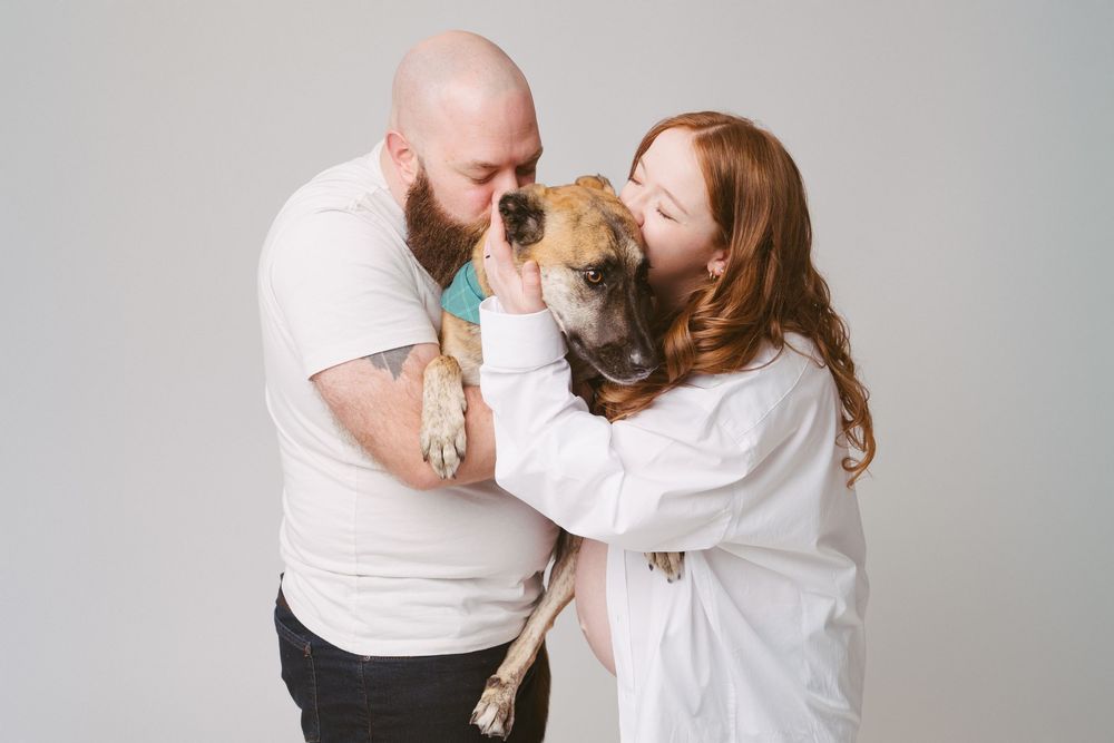 A bald man and redhead woman in white cuddle and kiss a brown boxer dog against a light gray background.