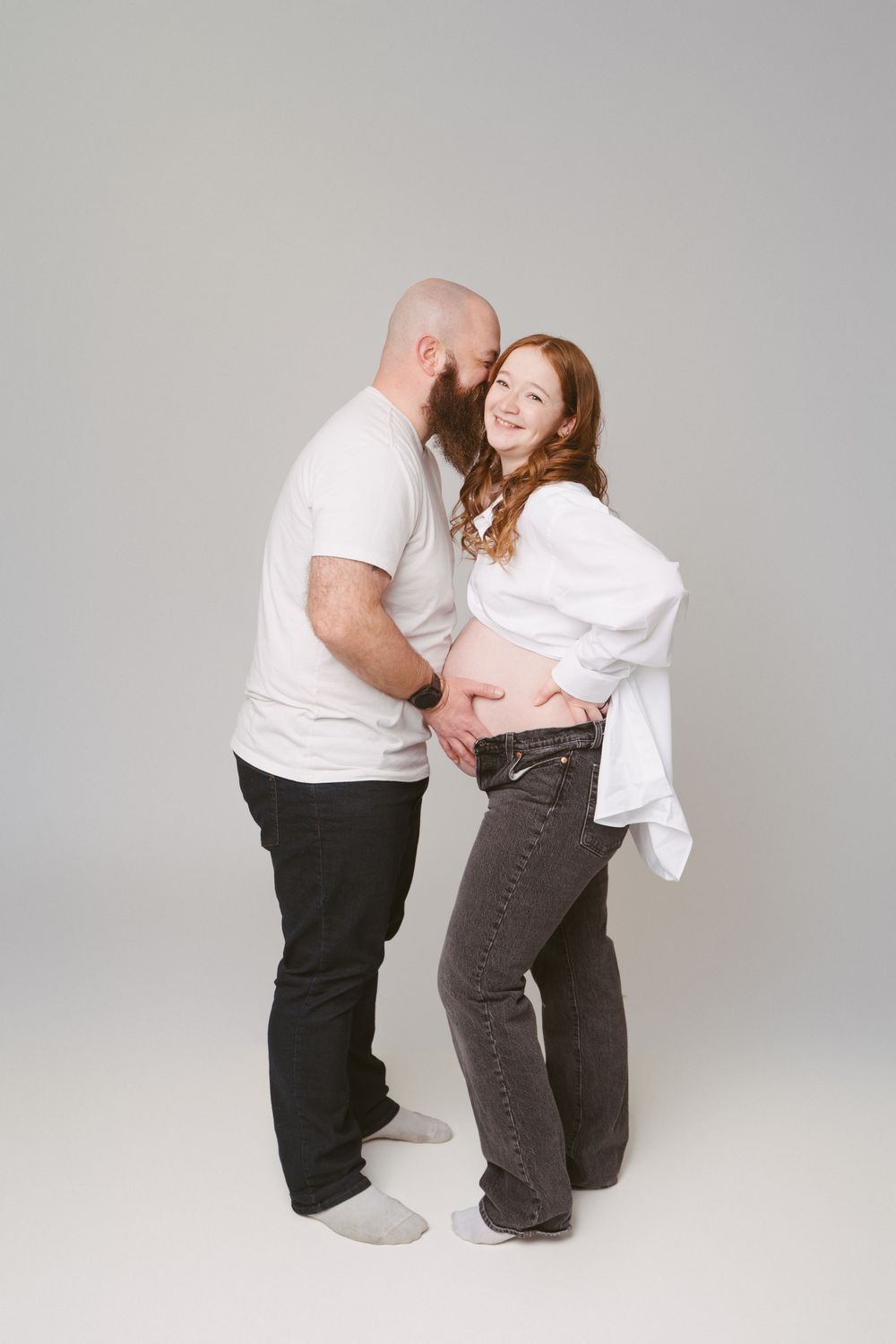 Bald bearded man embracing smiling pregnant redhead woman in white shirt and jeans in a studio setting.