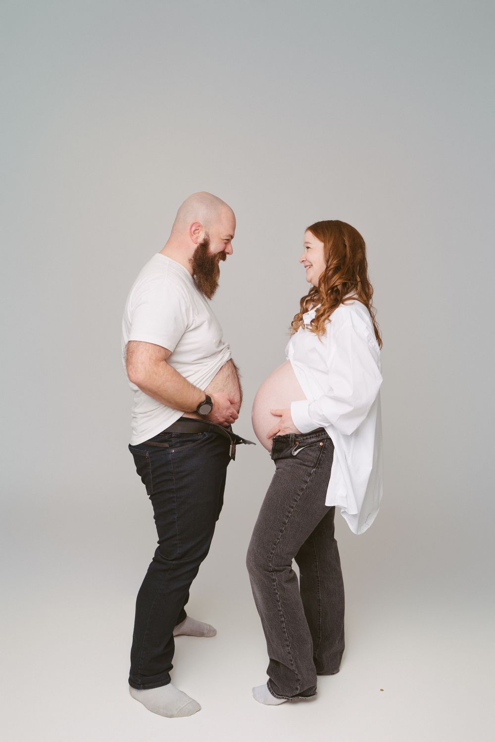 Pregnant couple laughing together, man lifting shirt to compare belly with his pregnant partner in a studio.