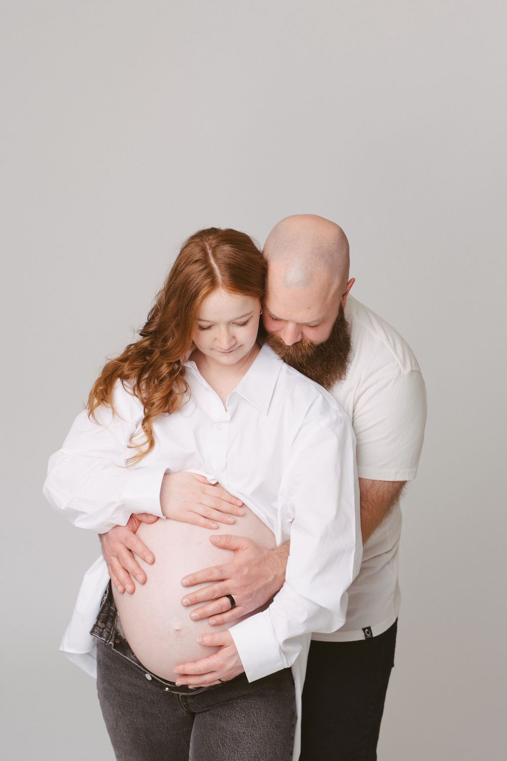 Pregnant woman in white shirt embraced by bearded bald man, both looking down at her baby bump in studio.