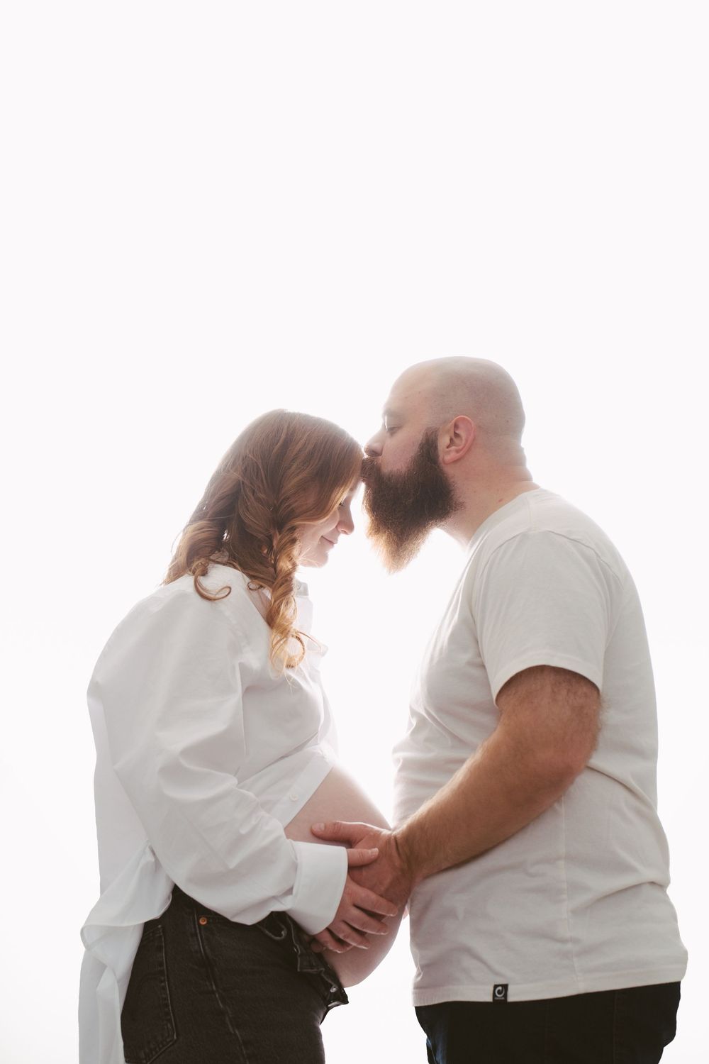 Pregnant couple holding baby bump together, bearded man kissing woman's forehead, backlit white background.