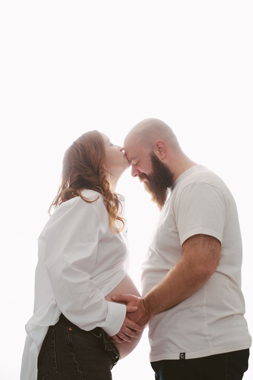 Pregnant couple holding baby bump and touching foreheads tenderly in a bright white backlit studio.