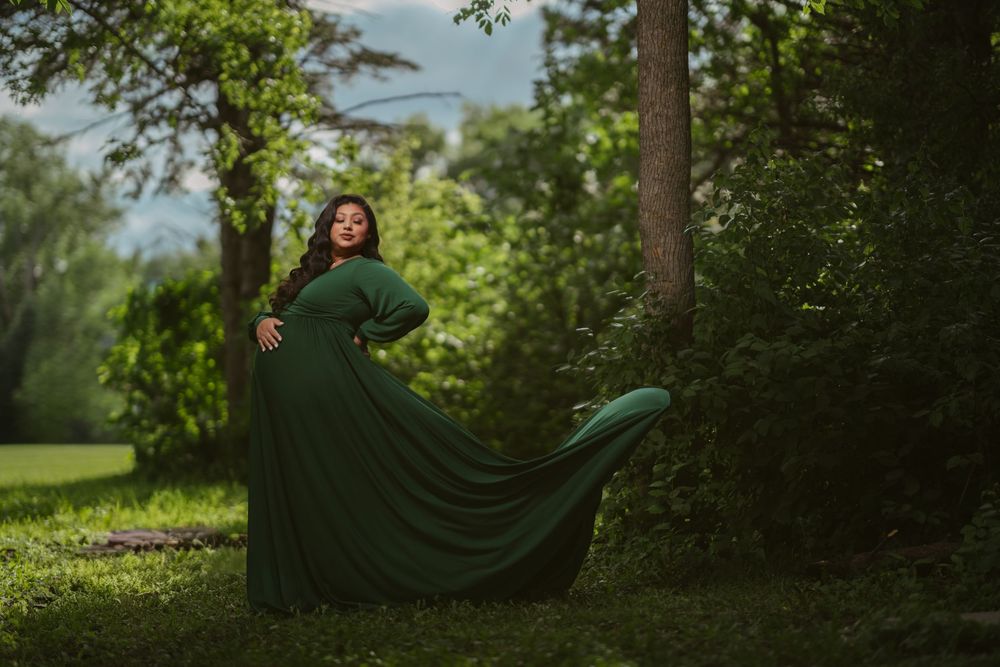 Woman in flowing green gown poses gracefully among lush trees in an outdoor maternity photography session.