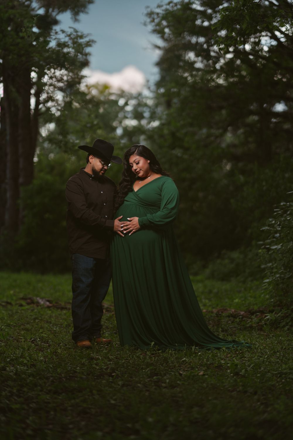Couple in green forest, pregnant woman in emerald gown, man in cowboy hat, maternity photo session outdoors.