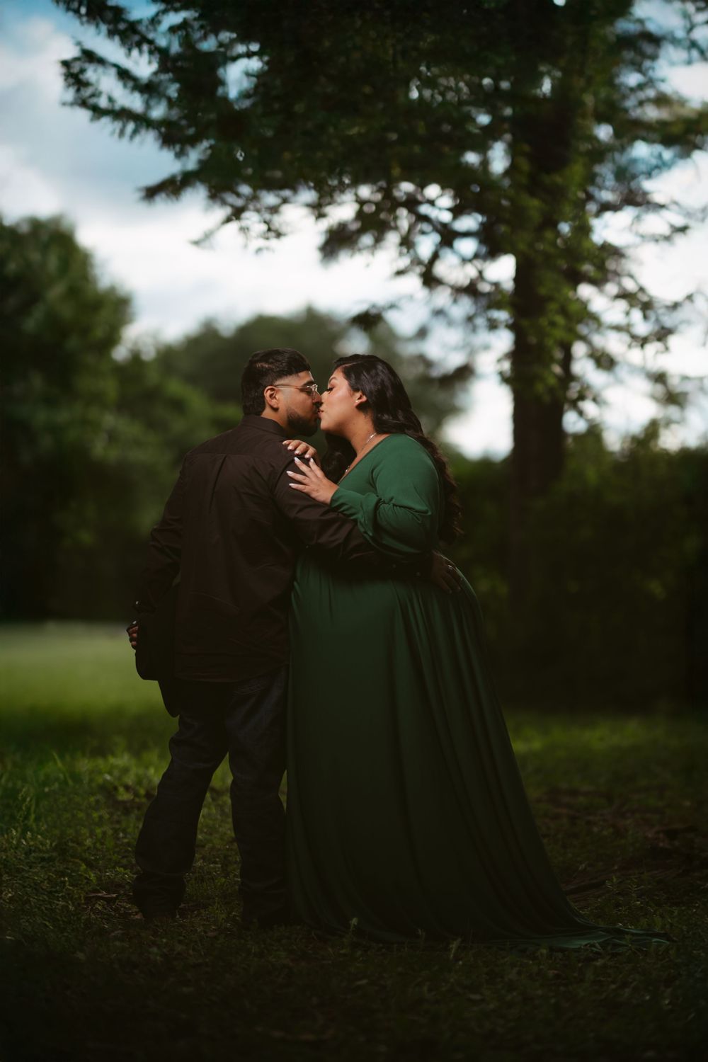 Couple sharing a romantic kiss outdoors, woman in green gown and man in dark suit, surrounded by lush trees.
