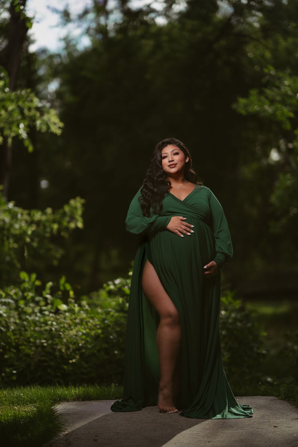 Pregnant woman in elegant emerald green maternity gown posing outdoors in lush green park setting.
