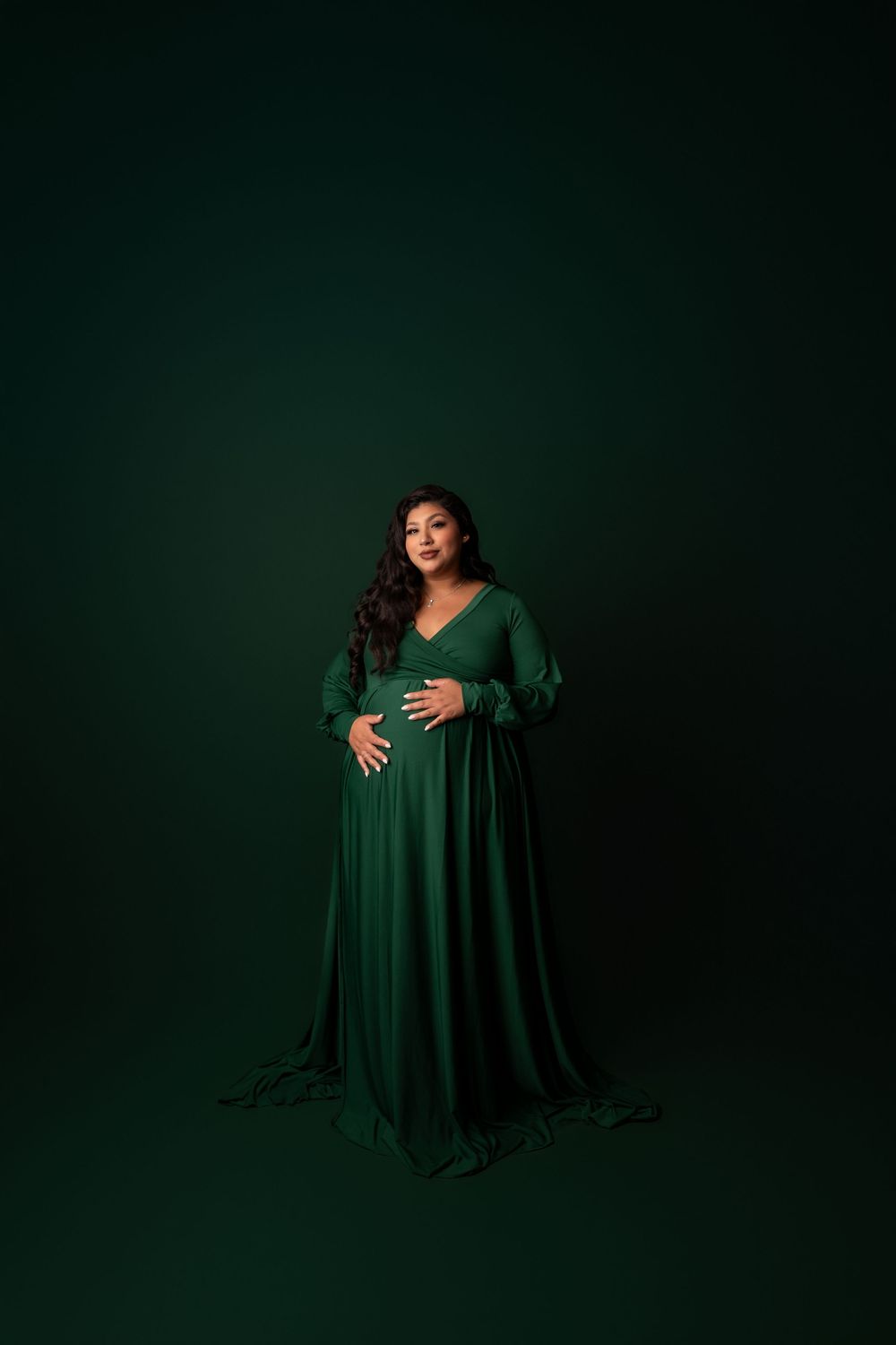 Pregnant woman in elegant emerald green maternity gown cradling her baby bump against a dark green studio background.