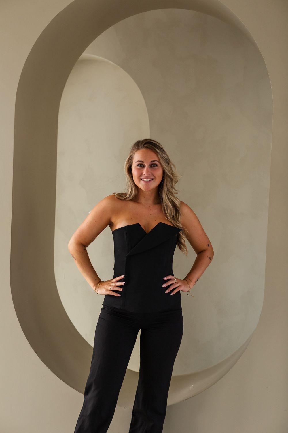 Model in black strapless jumpsuit strikes dynamic poses in front of curved white wall.