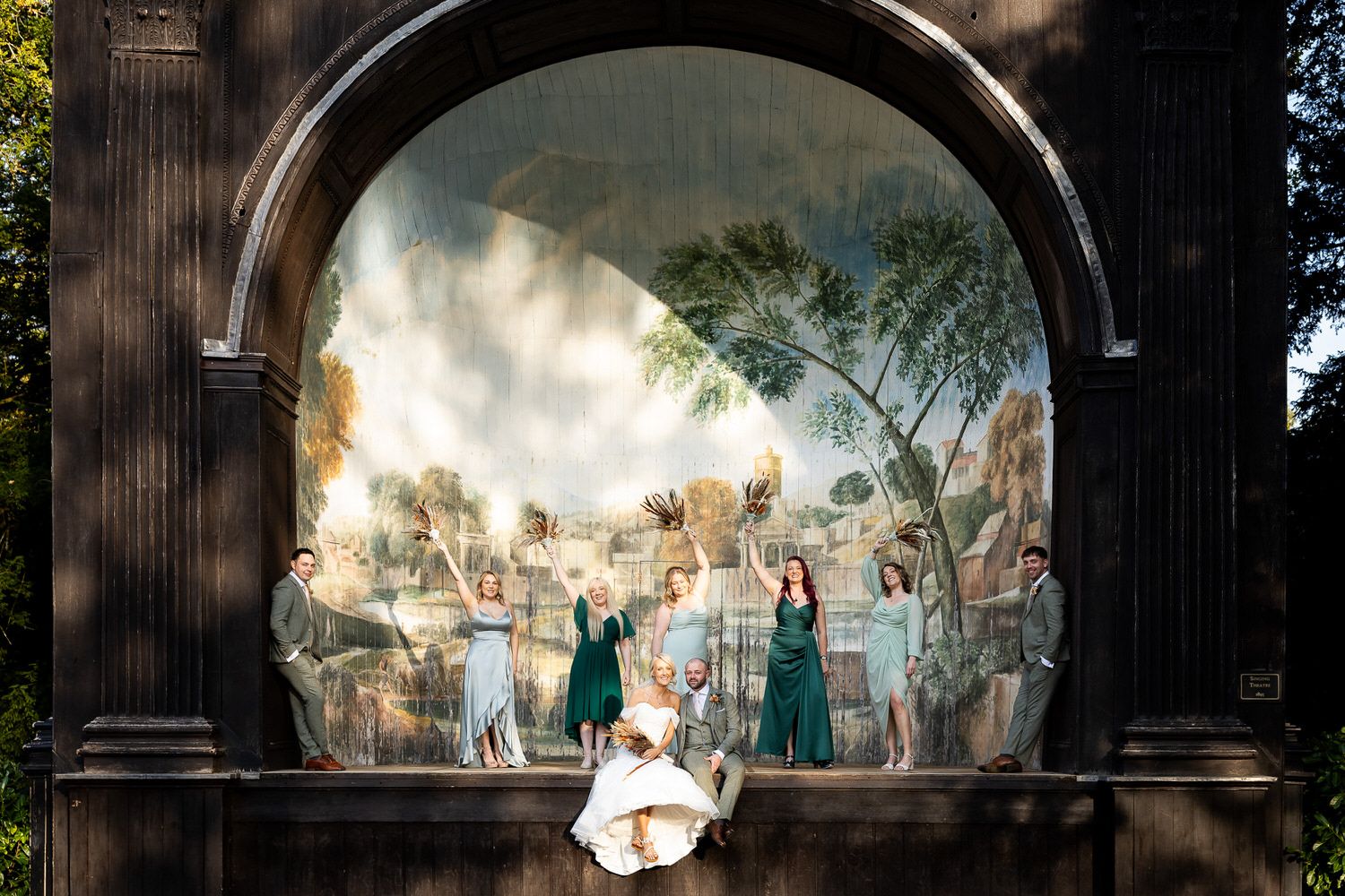 Wedding party posing in ornate arched alcove with scenic mural backdrop, bride and groom in foreground.