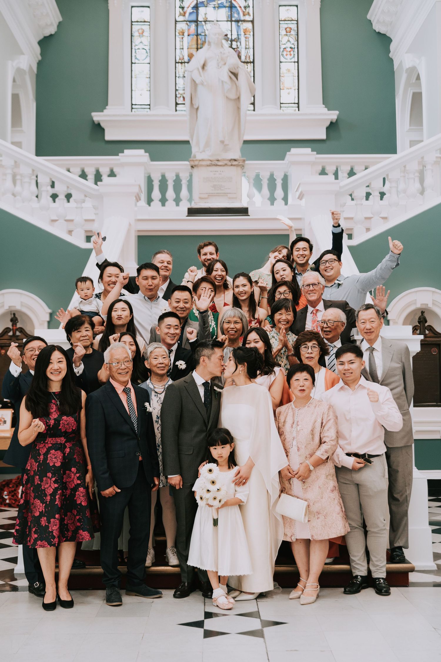 Bride and groom kiss surrounded by joyful wedding guests on the grand Woolwich Town Hall steps with a statue backdrop.