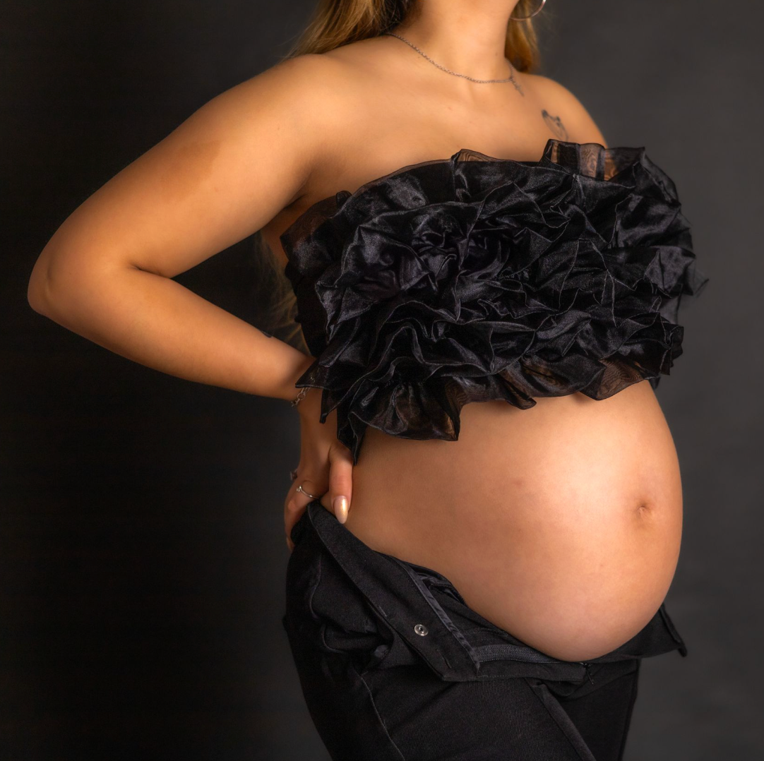Pregnant woman in black bikini cradling her baby bump while walking barefoot on a sunny sandy beach.