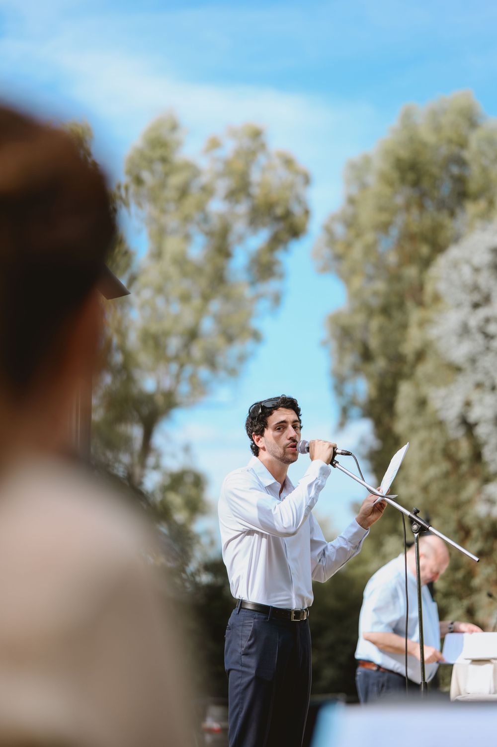 Man in white shirt speaking at microphone with paper at an outdoor wedding ceremony surrounded by trees.
