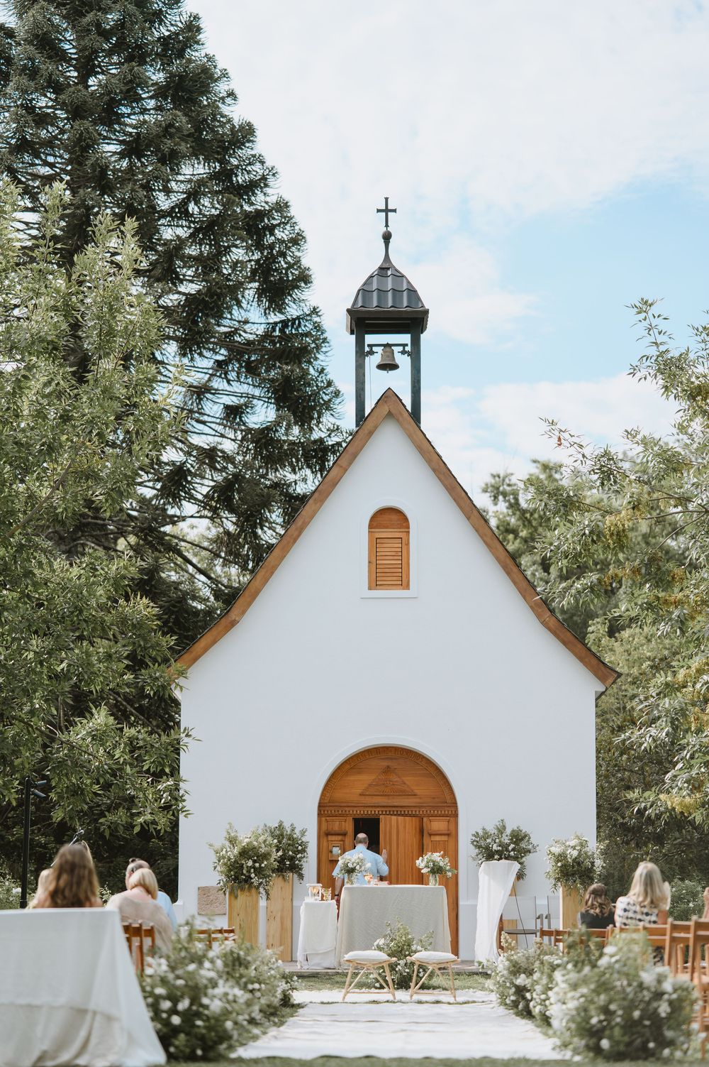 Charming white chapel with wooden cross and bell tower surrounded by lush trees at a wedding venue.
