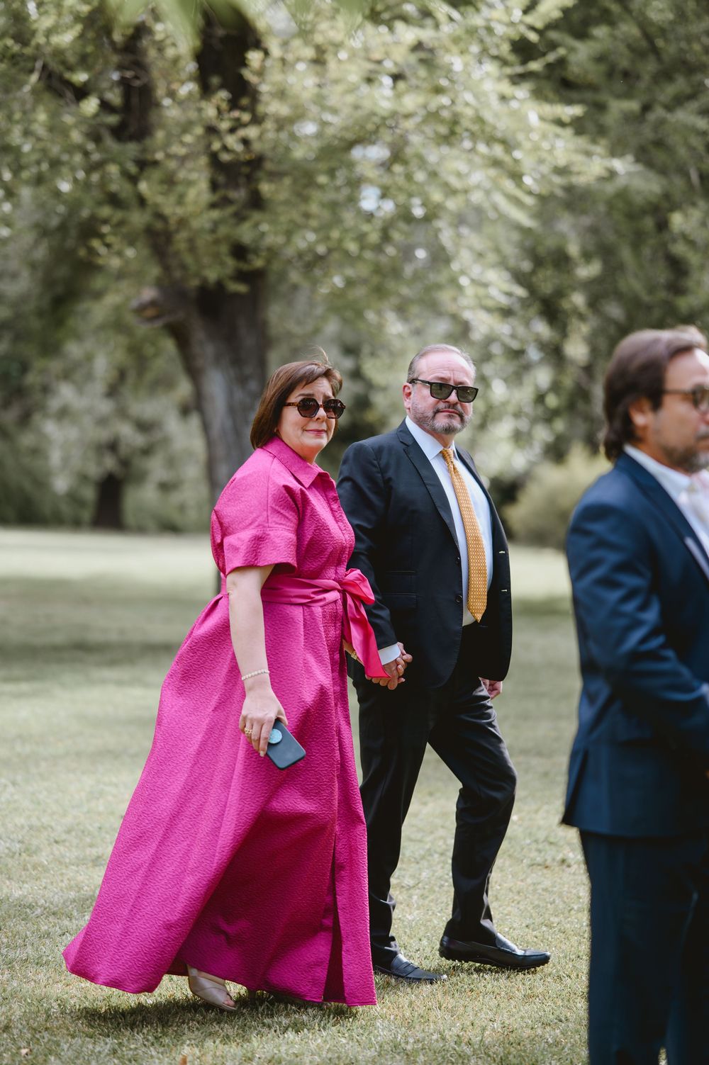 Elegant wedding guests walk outdoors, woman in bold pink maxi dress with man in dark suit and gold tie.