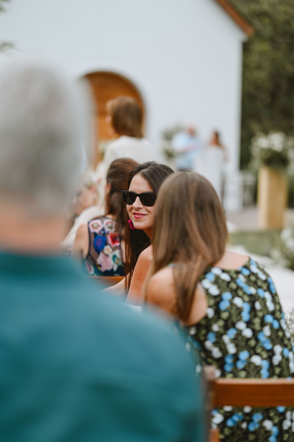 Wedding guests seated outdoors, smiling woman in sunglasses enjoys the ceremony on a sunny day.