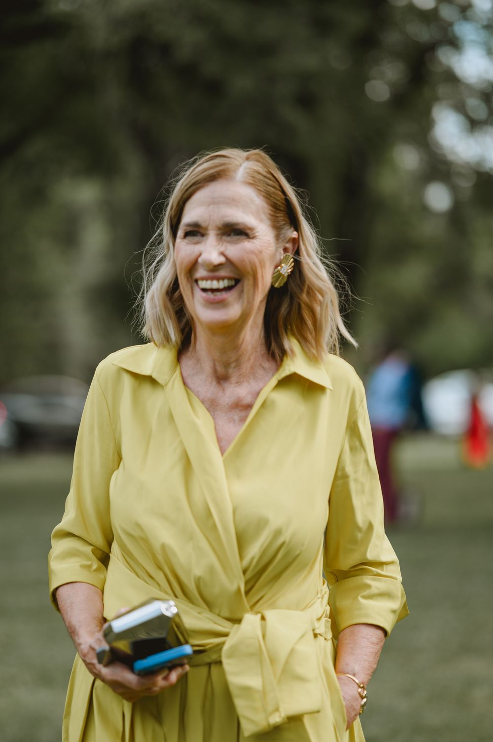 Smiling woman in yellow wrap dress holds phone while standing in green park at outdoor wedding.