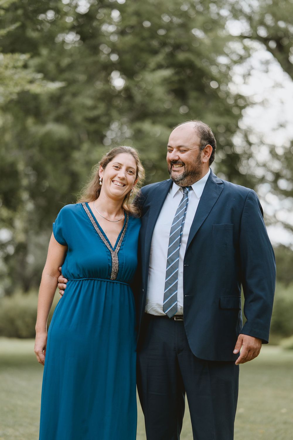 Smiling couple poses outdoors, woman in teal gown and man in navy suit with striped tie.