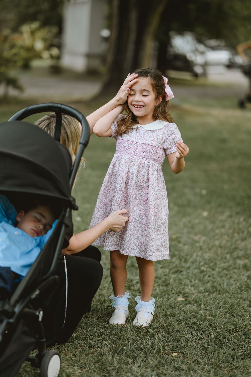 Adorable little girl in pink floral dress laughs while standing on grass near a stroller.