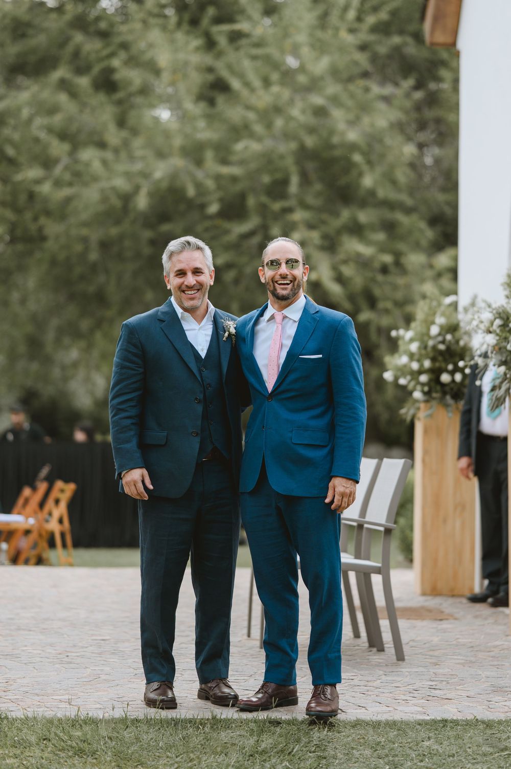Two men in navy suits smile together outdoors at a wedding ceremony venue.