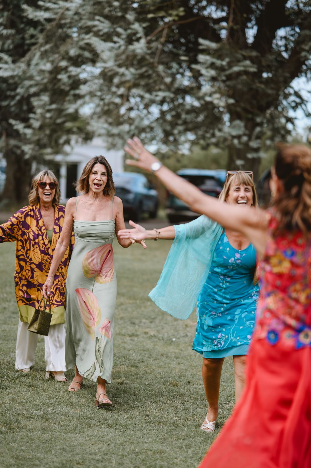 Group of cheerful women in colorful dresses greet each other with open arms on green lawn.