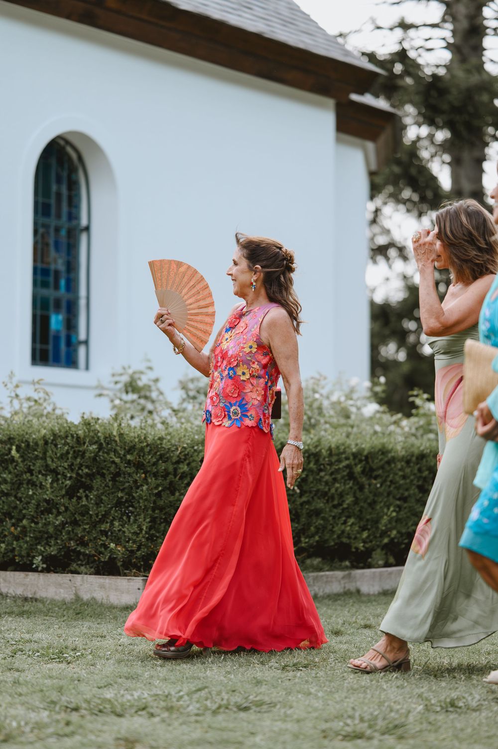 Woman in vibrant floral top and red skirt holds a fan outside a white chapel at a wedding.