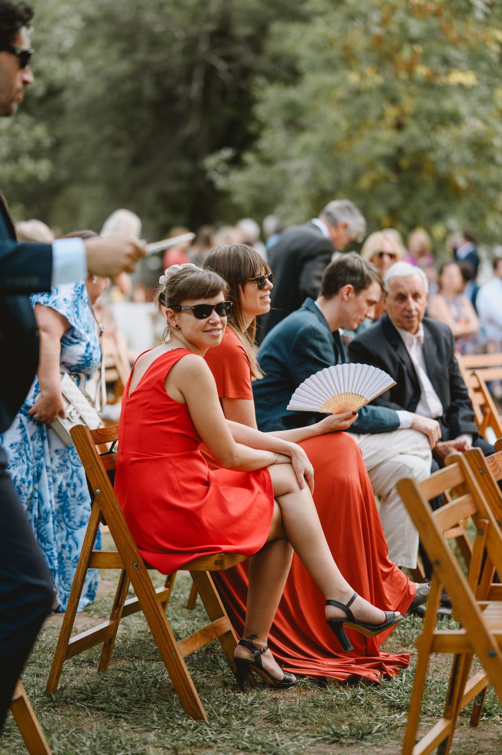 Wedding guests seated in wooden chairs outdoors, women in red dresses using fans on a warm day.