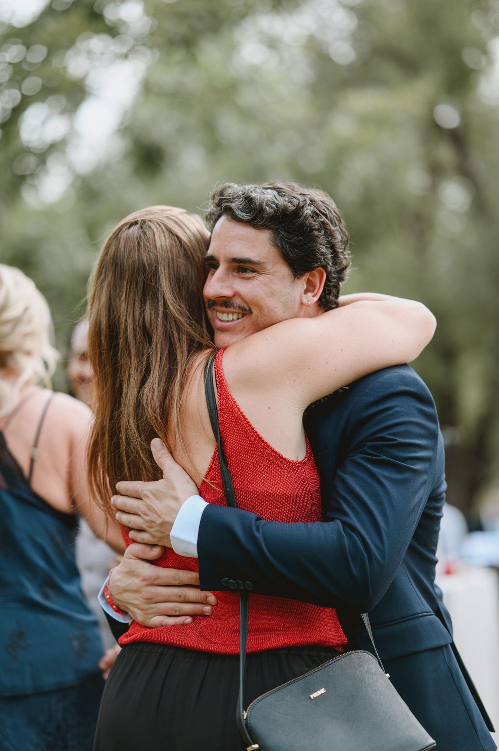 Smiling young man in navy blazer warmly embraces a woman in red at an outdoor wedding.