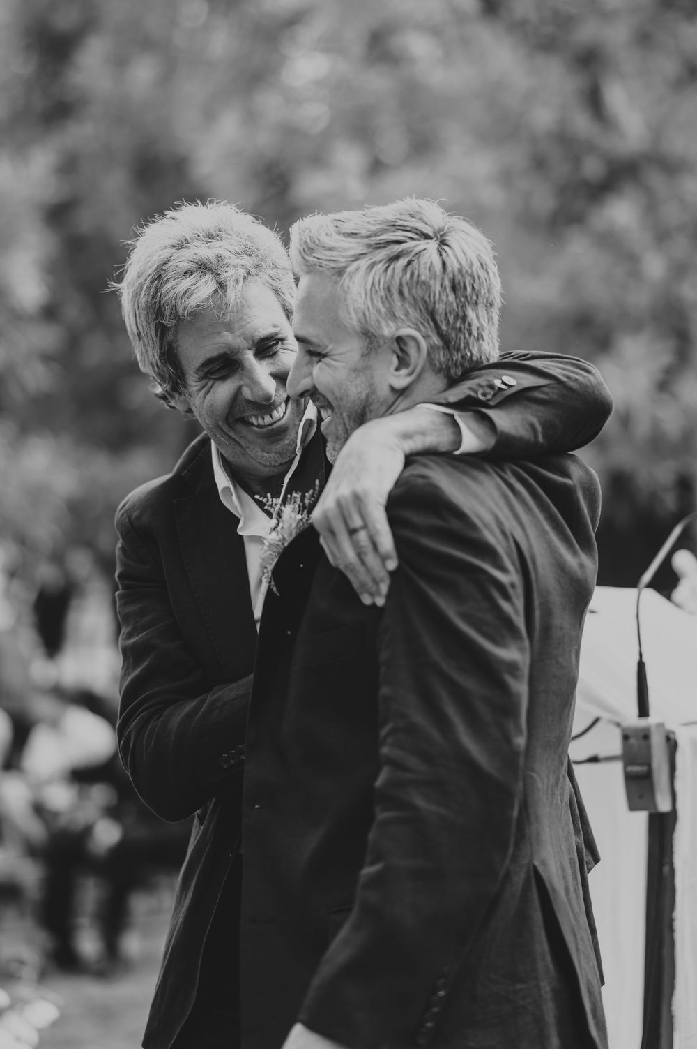 Two men in suits laughing and embracing at outdoor wedding ceremony, black and white.