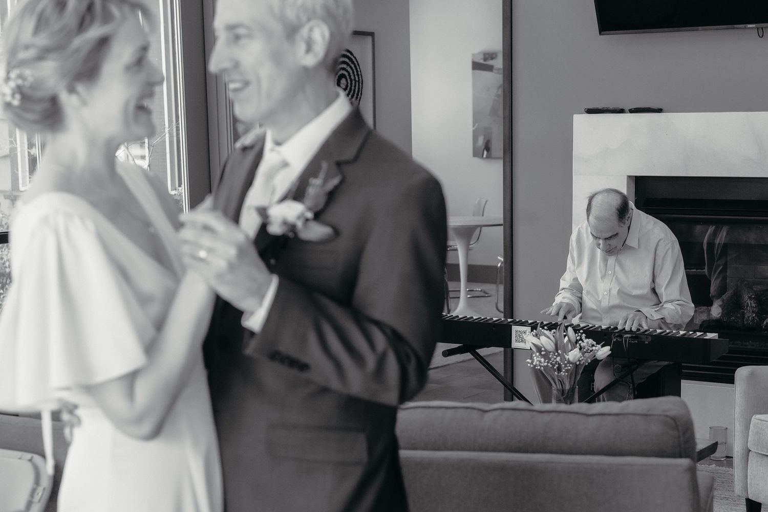 Black and white photo of a couple sharing a first dance at their wedding while a musician plays keyboard in the background.