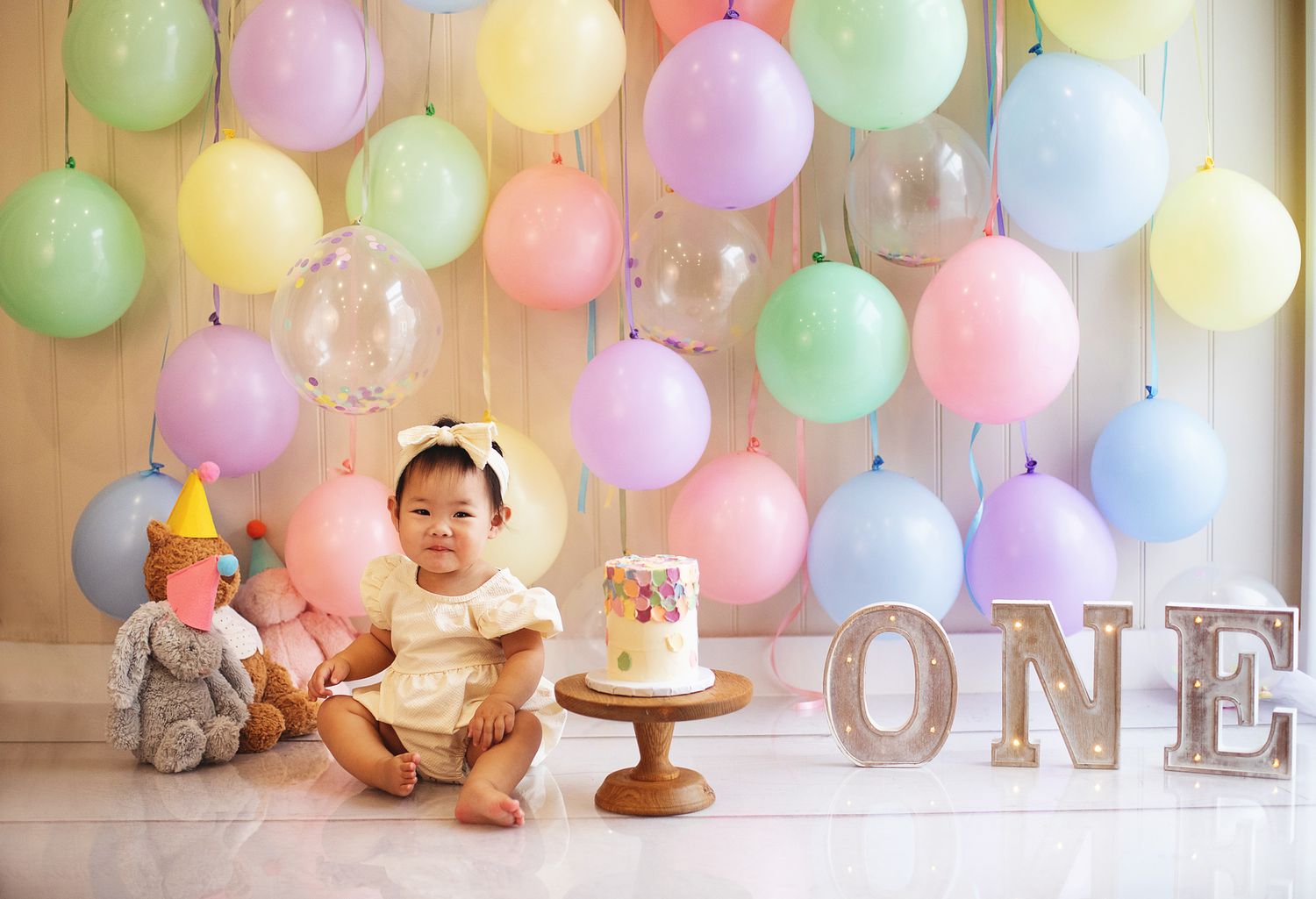 Baby girl in yellow dress sits before a first birthday cake, colourful pastel balloons, and ONE sign. During a cake smash session in Bexley