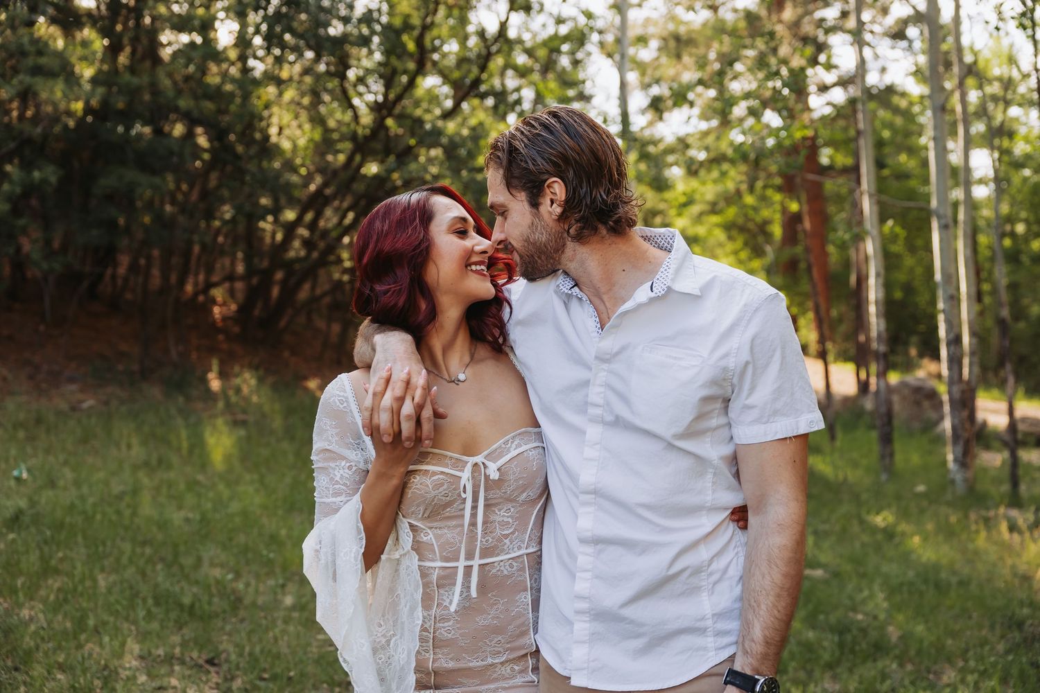 Couple sharing a romantic kiss outdoors, holding hands with an engagement ring visible, surrounded by lush greenery.