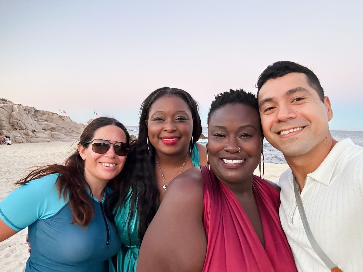 Four friends smiling together on a sunny beach, posing for a selfie with cliffs and ocean in the background.