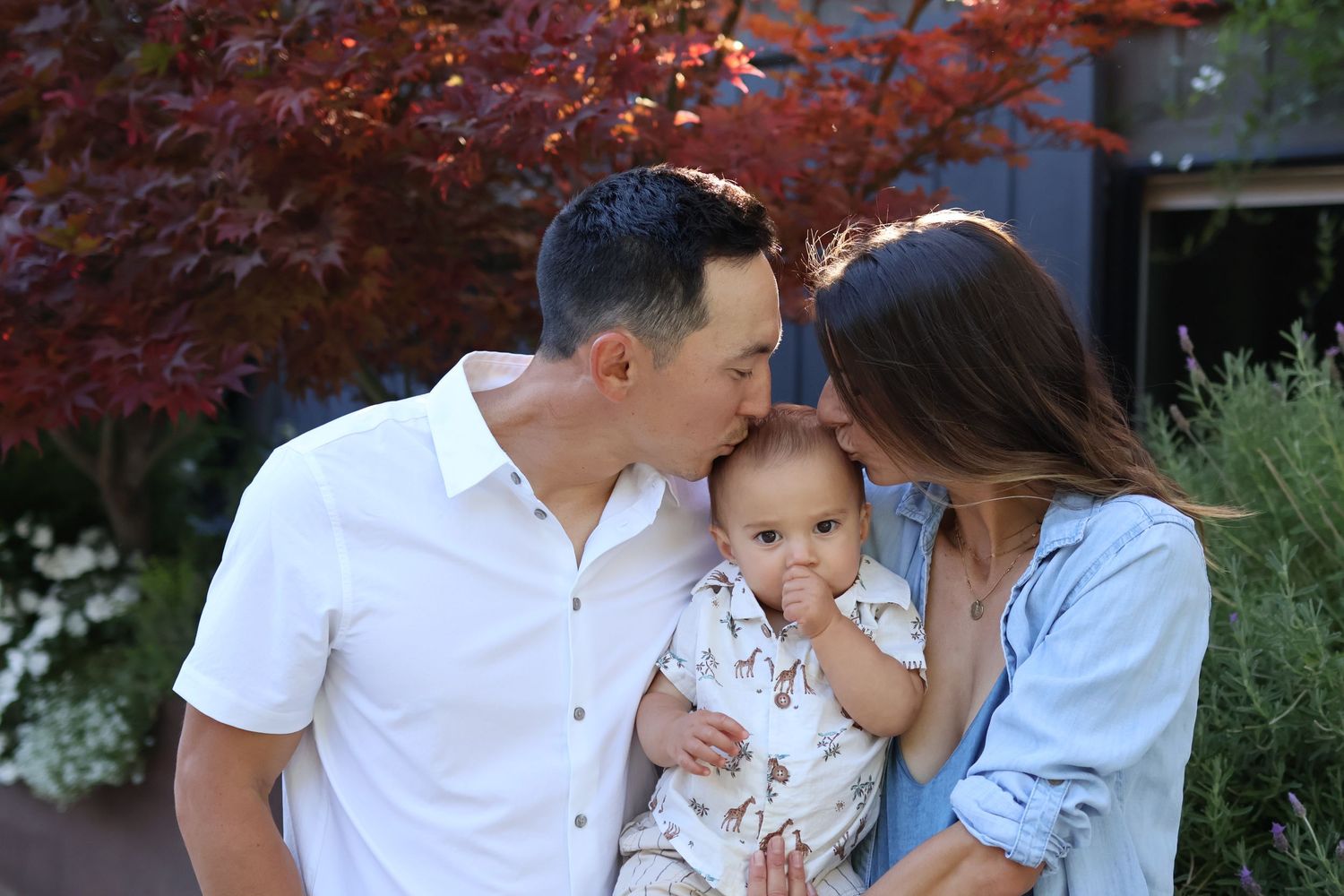 Mom and dad holding baby with fall tree in the background, both kissing babies head
